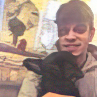 A person is smiling while holding a black dog. The background appears to be indoors with a window and some indistinct objects.